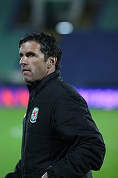 A man wearing a dark suit and white shirt standing at the edge of a football pitch