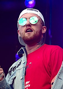 A tattooed Caucasian man holding a microphone smiles while rapping.