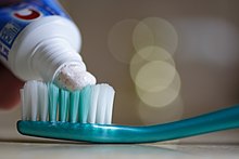 Foto mit verschwommenem Hintergrund; im Vordergrund wird Zahnpasta aus einer Zahnpastatube auf die Borsten einer Zahnbürste aufgetragen