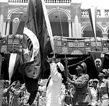 Photographie d'un homme en uniforme militaire hissant un drapeau au sommet d'un mat. Plusieurs personnes en uniforme se tiennent derrière lui.