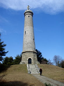 A very tall stone tower with a statue of a man at the top. It stands on a steep, grassy hill. A pathway and stairs lead to a door on the front of the tower. A person is walking along the pathway away from the tower.