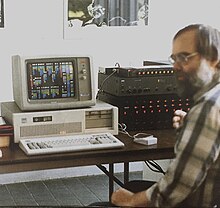 Фотография мужчины с бородой в клетчатой рубашке, сидящего за настольным компьютером рядом с устройством для микширования звука.