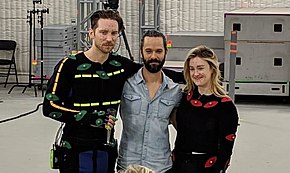 A man with slicked, dark hair holding a wine glass and wearing a black suit with motion capture dots on the suit and his face. He has his arm around a smiling man with dark hair and a dark beard. Next to him is a blonde woman, also in a motion capture suit with dots.