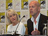 Bruce Willis speaks at San Diego Comic-Con. Actress Helen Mirren is seated to his right wearing a white shirt with the name Harvey Pekar