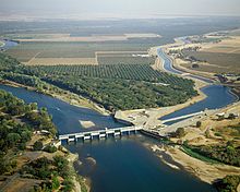 View from the air of a river flowing through the center from right to left; a dam blocks the water flow in the right half presumably to send some of the water to a canal that leads off to the upper left.