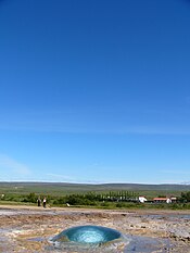 Circle of water in the ground with houses and a green skyline in the distant background.