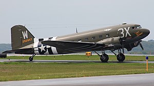 A drab green 1940s aircraft with "3X" painted in white near the cockpit and "W" on its tail faces right on a runway