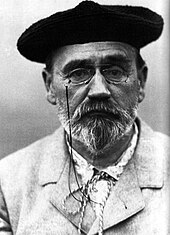 Photographie en noir et blanc d’Émile Zola montrant un homme déjà âgé, à moustache et barbe en pointe poivre et sel, le col noué d’une cordelette, portant des lorgnons et un béret de laine noire.