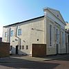 Three-quarter view of a wide, long stuccoed building, with the side wall more prominent. This is mostly a blank wall, but there are three windows towards the rear. The façade has three tall windows tapering from bottom to top; the central window has a small pediment and is shorter than the others to accommodate an entrance door. Above the roofline is a large pediment with louvred circular opening.