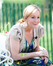 Seitliche Farbfotografie einer Frau, die auf einem weißen Stuhl vorgebeugt sitzt. Sie hat ihre blonden Haare zusammengebunden und trägt eine kurzärmelige, graue Weste über einem blau-grünen Kleid. An ihrer linken Brust ist ein Ansteckmikrofon.