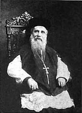 An elderly man with a long beard, a hat, and a crucifix hanging from his neck is sitting on an ornate chair.