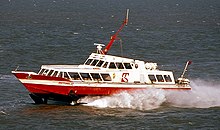 Broadside view of hydrofoil, with red hull and white superstructure, travelling right to left