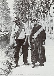 Reproduction noir et blanc d’une carte postale du début du siècle représentant un couple de haleurs sur le chemin de halage. Un vieil homme à gauche et une femme âgée à droite. Chacun porte une sorte de sangle appelée « las », en travers de son torse, reliée par une corde à l'embarcation à gauche de l’image.