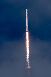 A white Falcon 9 rocket cuts through the blue sky, with its nine engines producing a bright yellow flame.