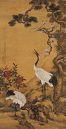 Two cranes near a pine tree. One is feeding on the ground while another rears its head high. Red flowers are also in the background
