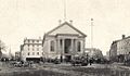 Black and white photo of a grand Greek revival civic building with a gable roof, an oversized portico supported by four large ionic columns flanked to the left and right by tall, barrel arch windows, below a simple pediment. The building has a public open space paved with dirt and granite pavers and crossed by streetcar tracks in front of it. The building is surrounded by nineteenth century mixed use urban buildings, mostly of brick and granite. There are multiple people and horse-drawn carts in the public space.