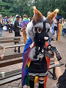 Das Foto zeigt im Vordergrund eine Person mit einer überdimensionierten schwarz-grau-hellbraunen Tiermaske, Pfotenhandschuhen, einer Regenbogenflagge als Umhang und schwarz-weißer Kleidung; im Hintergrund sind zwei Parkbänke sowie viele Personen zu sehen.