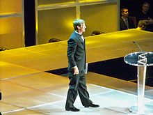 Un homme est sur scène habillé en smoking et s'approche d'un pupitre en tenant son discours dans la main gauche.
