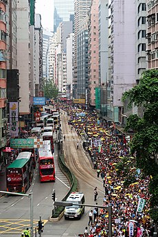 a large procession along the east-bound carriageway of a road through a built-up area; severe traffic congestion in the westbound carriageway