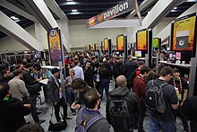 Des joueurs font la queue devant différents stands de jeux vidéo oranges.