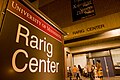 A closeup of a sign on the left, the top text reading "University of Minnesota", and the bottom, slightly larger text reading "Rarig Center". A building's exterior on the right, made up of concrete and brick has large doors under an overhang, and windows with their blinds slightly drawn. In front of the building, people are standing around and talking.