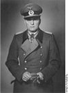 A black-and-white photograph of a man wearing a military uniform and coat, peaked cap holding gloves in his hands.