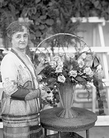 A woman with gray curly hair looking at the camera to the viewer's left of a flower bouquet.