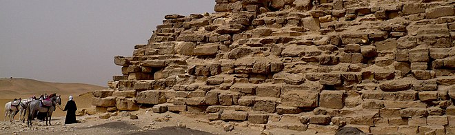 Photo panoramique au pied d'une pyramide, des cavaliers vus de dos à gauche.