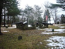 A monument featuring four black cannon barrels mounted on a stone wall in the middle of a small cemetery. The ground is partly covered with snow. Many trees stand in the background. The sky is cloudy.