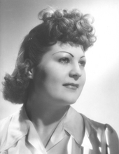 Photo en noir et blanc d'une femme d'une cinquantaine d'années, les cheveux bouclés et en chignon sur le front, posant en esquissant un très léger sourire.