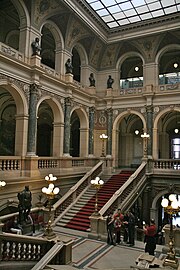 Vue intérieure d'un édifice imposant : il s'agit d'un grand escalier, au centre d'un grand hall. Les deux étages visibles sont constitués de couloirs à colonnade ouverts sur le hall.