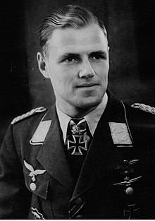 A black and white photo of the head and shoulders of a young man. He wears a peaked cap and military uniform with an Iron Cross displayed at the front of his shirt collar.