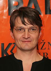 Detail obličeje Jana Svěráka stojícího před reklamním billboardem oranžové barvy