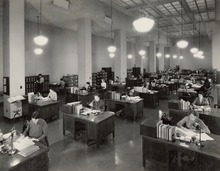 Photographie noir/blanc présentant des personnes assises à des tables alignées dans un grand bureau ouvert.