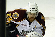 A Caucasian ice hockey player bent over at the waist. He wears a white helmet and has a large beard.