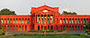 A wide red building, with numerous pillars and archways, stands in the middle of some gardens. A road runs just in front of the court.