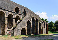 A photo, taken at a distance, of the exterior stairs of the Pompéi amphitheatre. The stairs form an inverted V besife the amphitheatre wall and contain several arches within it for access