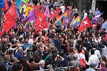 Grupo de pessoas com bandeiras coloridas com o símbolo do Bloco de Esquerda e o símbolo feminista. Nesta fotografia estão presentes alguns membros do partido como Catarina Martins, Mariana Mortágua, José Soeiro, Marisa Matias, Pedro Filipe Soares. Também estão presentes jornalistas e operadores de câmara.