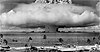 Bikini Atoll nuclear test, a wide mushroom cloud spreads out above an atoll, surrounded by moored ships with a sandy beach and palm trees in the foreground