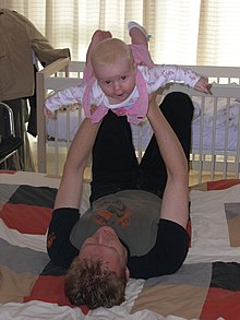 Un jeune père allongé sur le dos sur un édredon sur le sol. Il tient sa fille bébé au-dessus de lui avec ses bras tendus et ses mains autour de sa cage thoracique. Le bébé a les bras et les jambes tendus et arque son dos, il sourit à l'appareil photo.