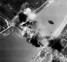 A black-and-white photograph showing an explosion on a railway bridge, with an American bomber flying overhead.