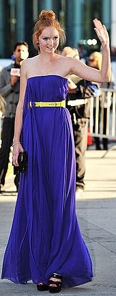 Cole outside wearing a strapless purple dress with her hair up in a large bun, surrounded by photographers.