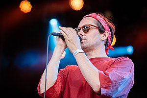 Cropped image of Milosh wearing a red t-shirt, red bandana, and sunglasses, standing onstage, appearing to sing into a microphone