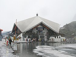 A stone building is surrounded by partially frozen ponds. Pilgrims can be seen on the paths