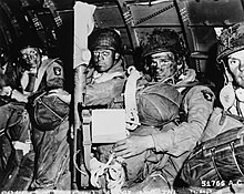 A black and white image of three men and the head of a fourth, all in combat gear and with their faces smeared with black, sitting against the metallic interior wall of a plane, and carrying heavy packs of gear. The soldier in the center foreground has a piece of paper in his left hand and holds a rocket launcher upright with his right arm.