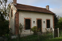 Photo couleurs de la maison éclusière du Hateau. Il s’agit d’une maisonnette à base rectangulaire. Les angles des murs sont constitués d’un chaînage en briquettes rouges. Il en est de même des deux fenêtres qui encadrent symétriquement la porte d’entrée en façade. La toiture est en tuiles et est bordée de deux cheminées en briques