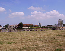 Landscape photo dominated by a field in front, surmounted by blue sky with white clouds. A row of modern buildings crosses the center; in front of the buildings a few stones can be seen piled atop each other.