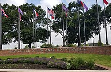 Photographie en couleurs d'un muret en brique où est inscrit le nom de l'université, sur lequel figurent plusieurs drapeaux aux couleurs des États-Unis, du Texas et de l'université.