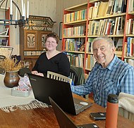 Två glada deltagare i skrivstuga sitter med varsin dator vid ett bord i ett rum pyntat med föremål från Hälsingland.