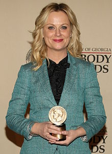 Amy Poehler faces forward wearing a light blue blazer. She holds a small circular award by its base.